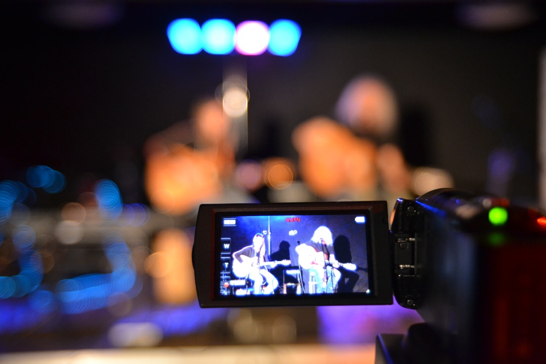 Im Bildvordergrund steht ein Display eines Videorecorders, auf dem zwei Personen zu sehen sind. Es sind zwei Gitarristen, die auf einer Bühne ein Konzert spielen.